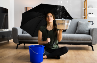 Eine Frau sitzt im Schneidersitz auf dem Boden in ihrem Wohnzimmer, mit einem Regenschirm in der Hand und einem Eimer neben sich stehen. Sie sieht traurig aus und es tropft über ihr von der Decke..